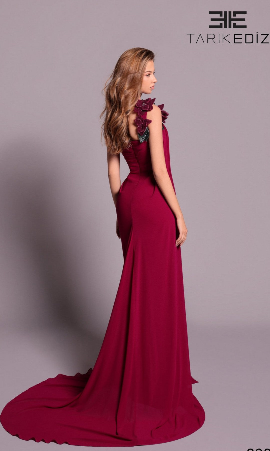 A woman models the Tarik Ediz 99036, a floor-length sleeveless magenta formal dress with 3D floral shoulder detail, standing on a plain background. The Tarik Ediz logo appears in the corner.